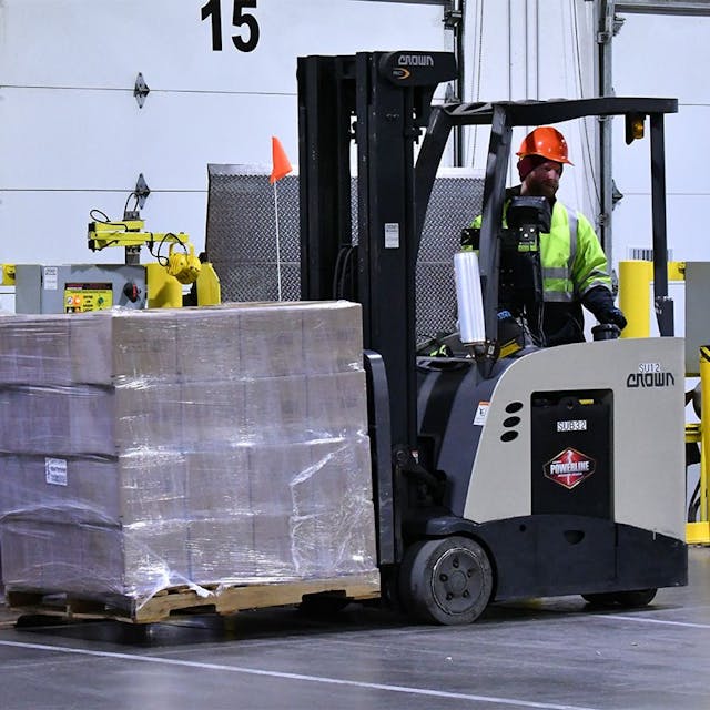 Man wearing hard hat driving a forklift with a pallet in front of a dock door.